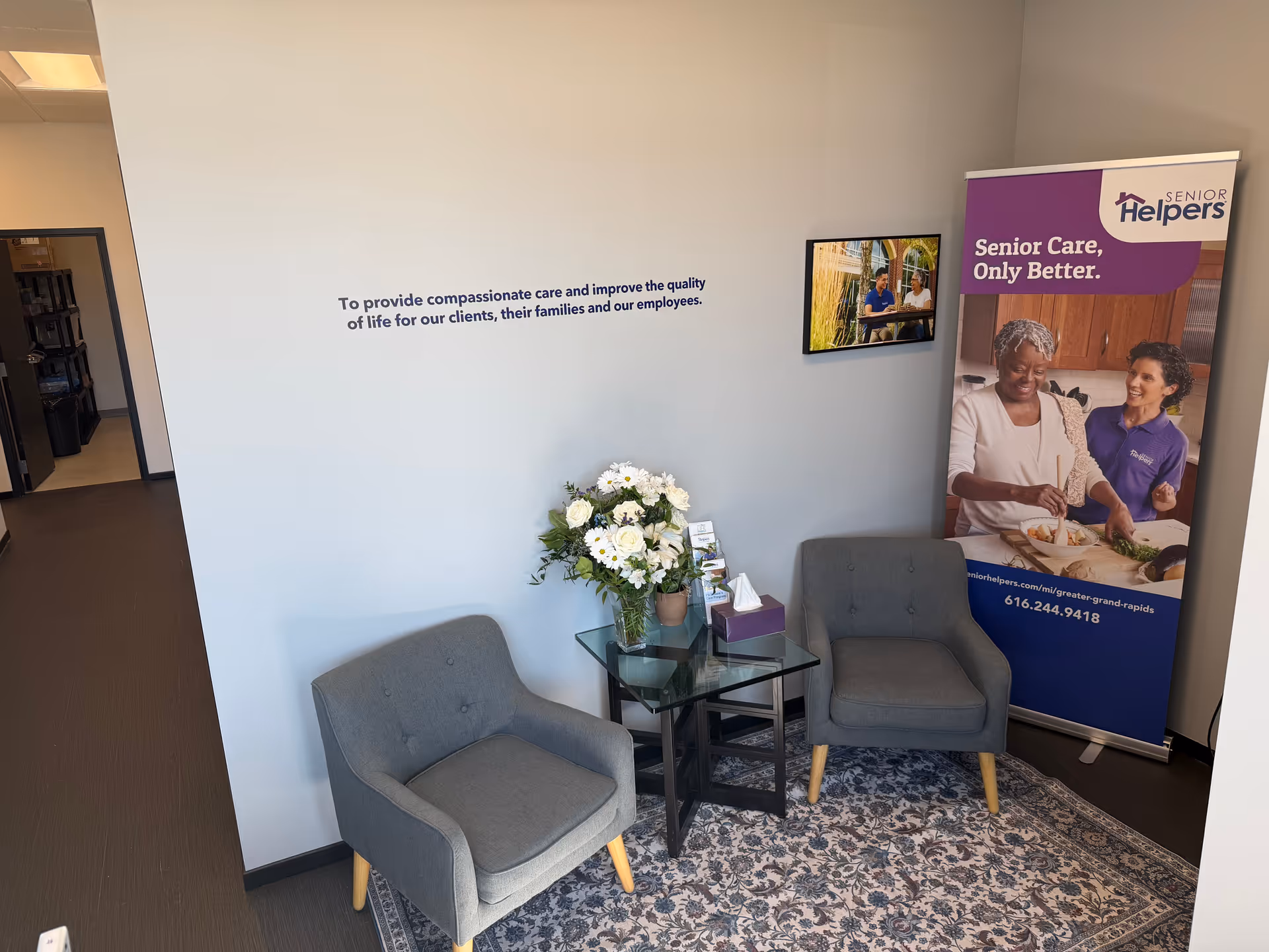 Two gray armchairs flank a glass table with flowers beside a Senior Helpers banner.