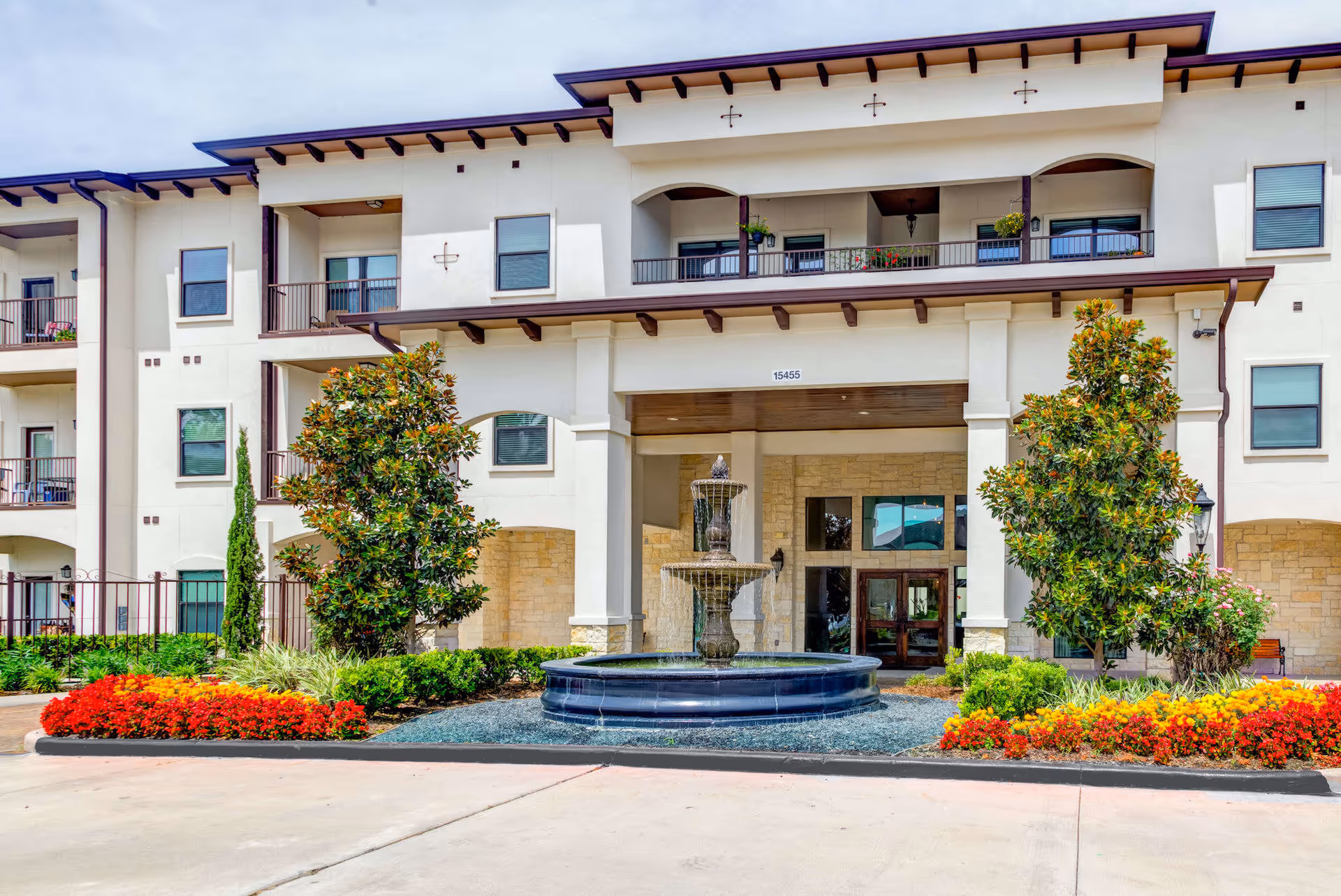 Front exterior view of The Laurel at Vintage Park building featuring a three-story structure with balconies, a central entrance with a covered portico, a decorative water fountain in front, and landscaped flower beds with red and yellow flowers and green shrubs.