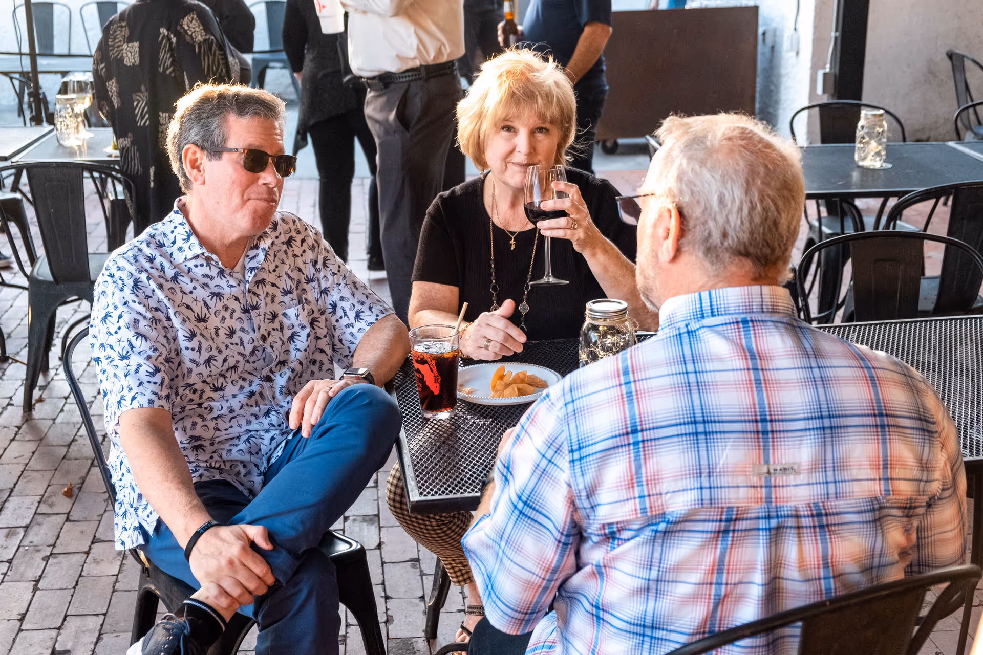 Three older adults seated at an outdoor patio table, enjoying drinks and snacks.