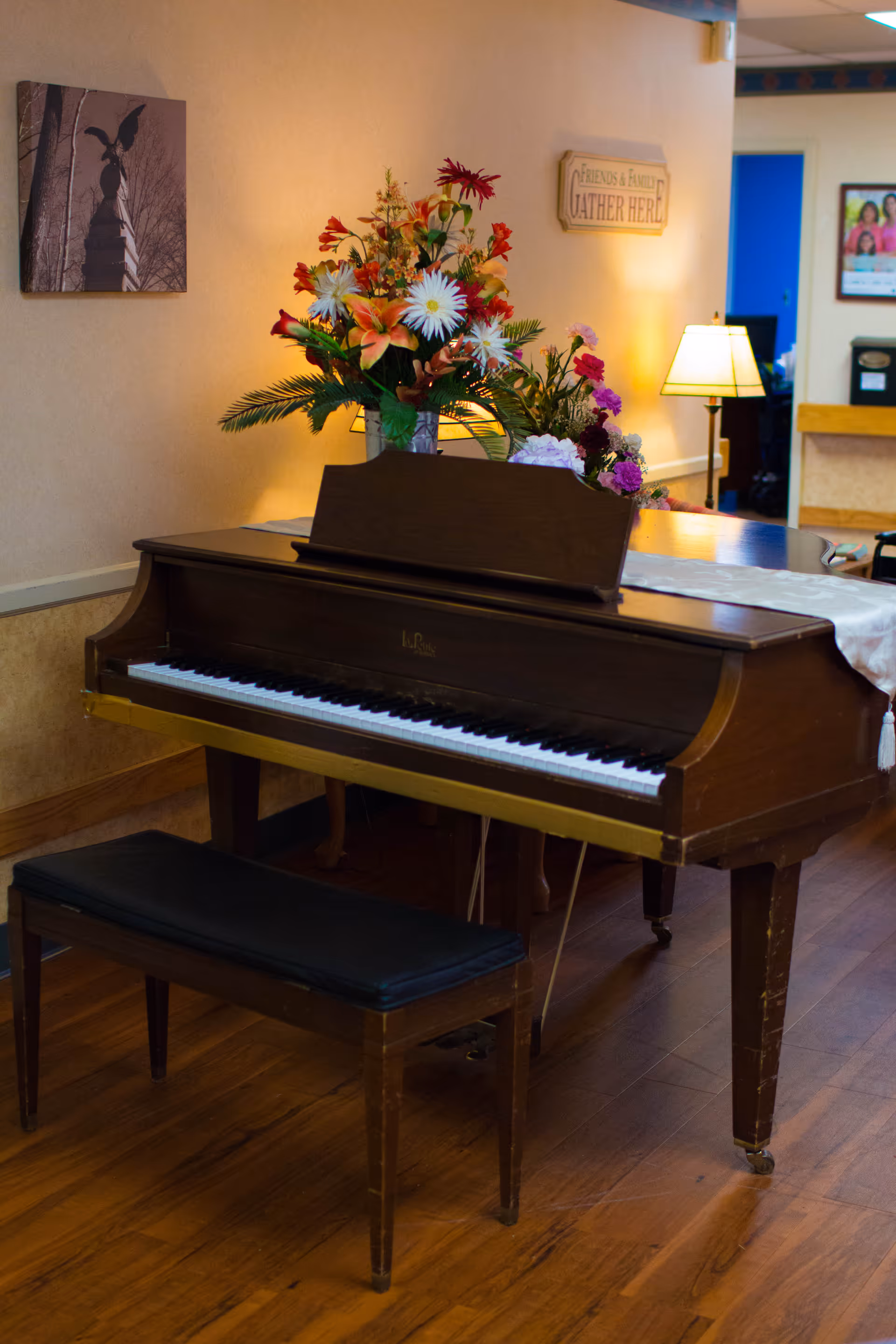 A wooden upright piano with a black cushioned bench in front of it, decorated with a large bouquet of colorful flowers on top. The room has wooden flooring, a lamp on a side table, a wall sign that reads 'Friends & Family Gather Here,' and a framed picture on the wall.