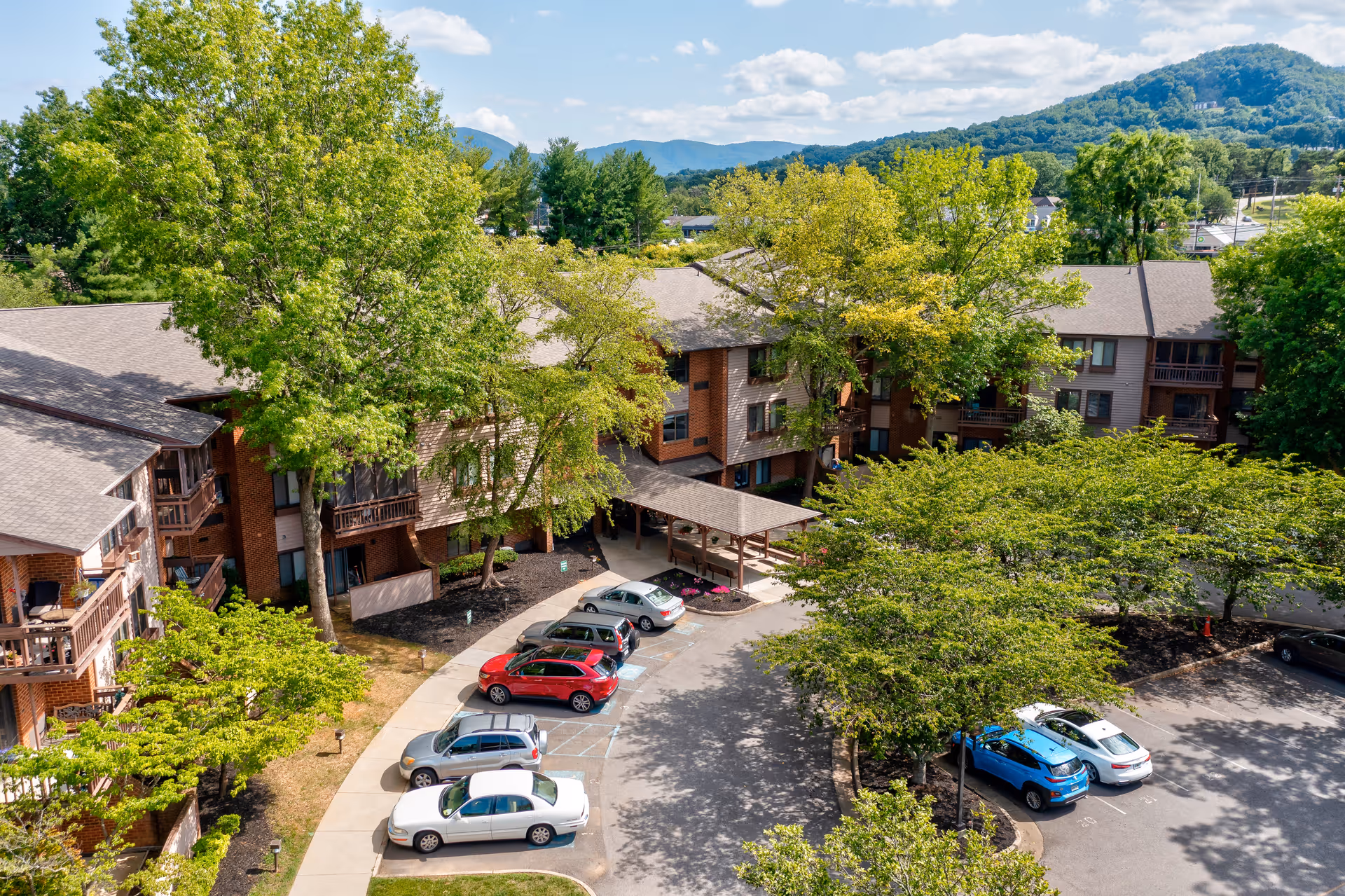 An exterior view of a brick senior living building surrounded by trees and parked cars.