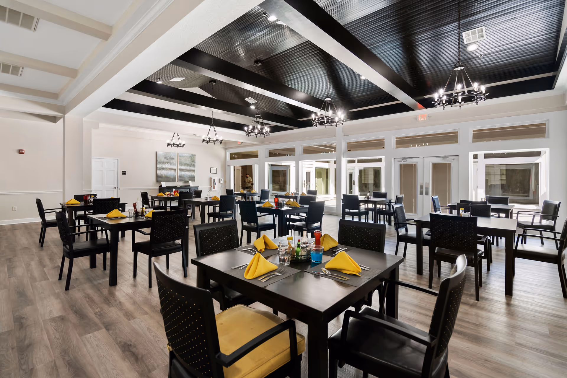 A spacious dining room with dark tables, yellow napkins, chandeliers, and large windows.