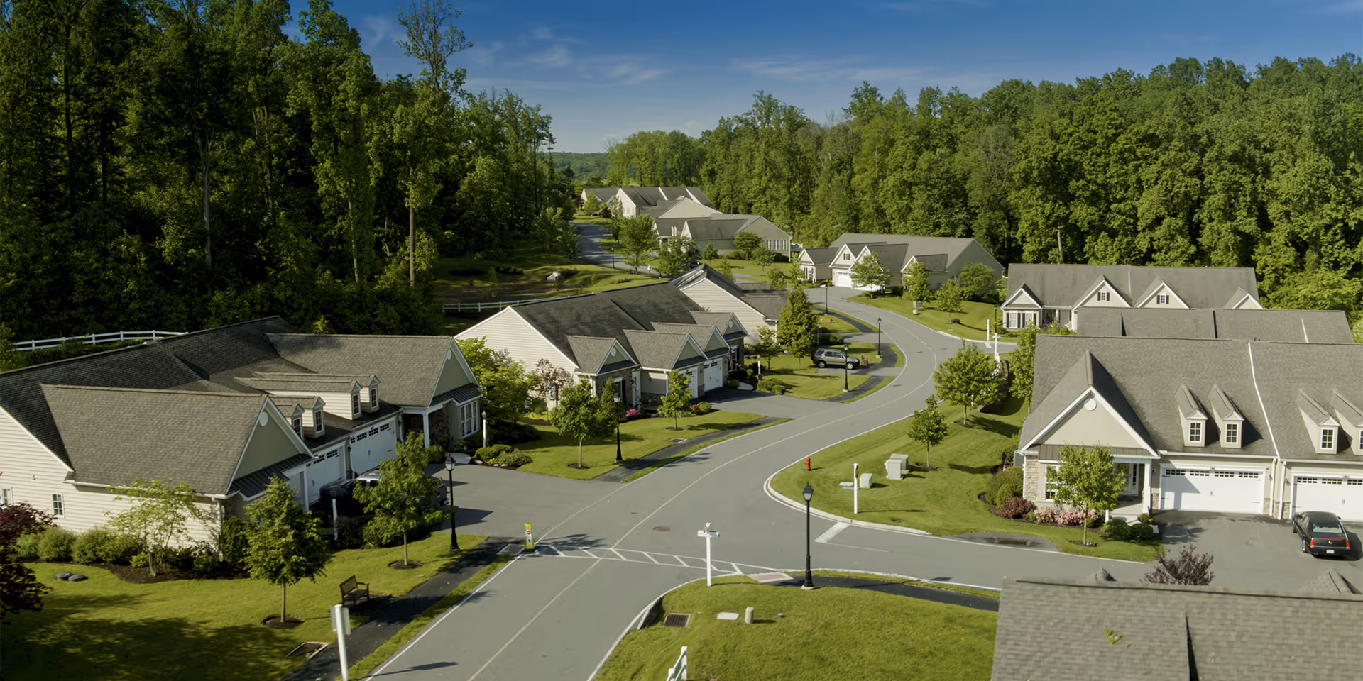 Aerial view of a residential retirement community with multiple single-story houses along a curved road, surrounded by green lawns and trees under a clear blue sky.