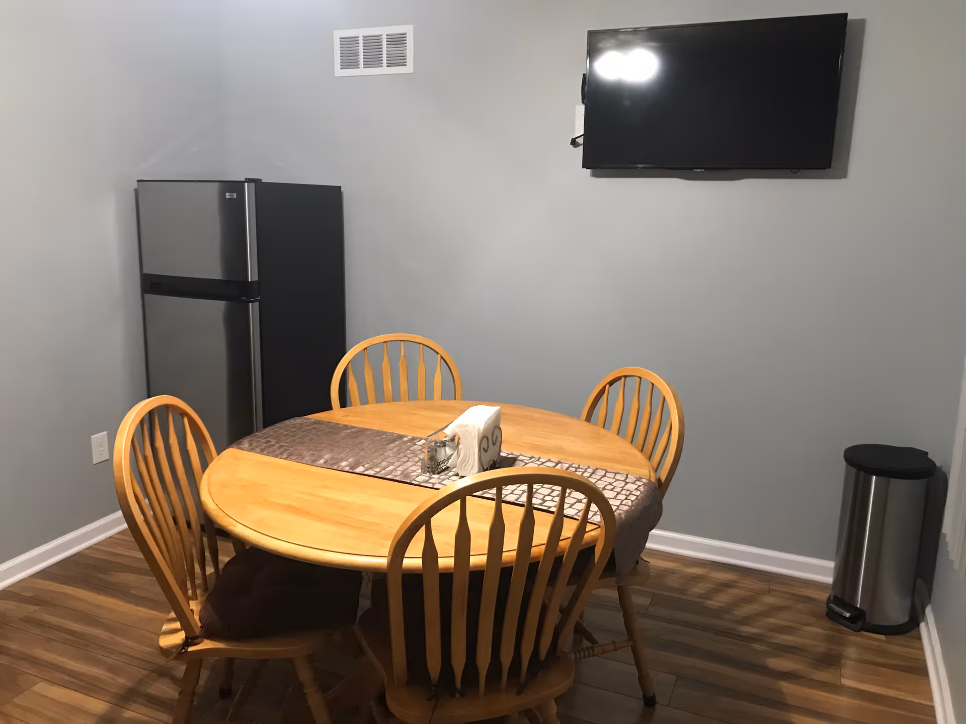 Round wooden table with six chairs in a small dining area; fridge and TV on wall.