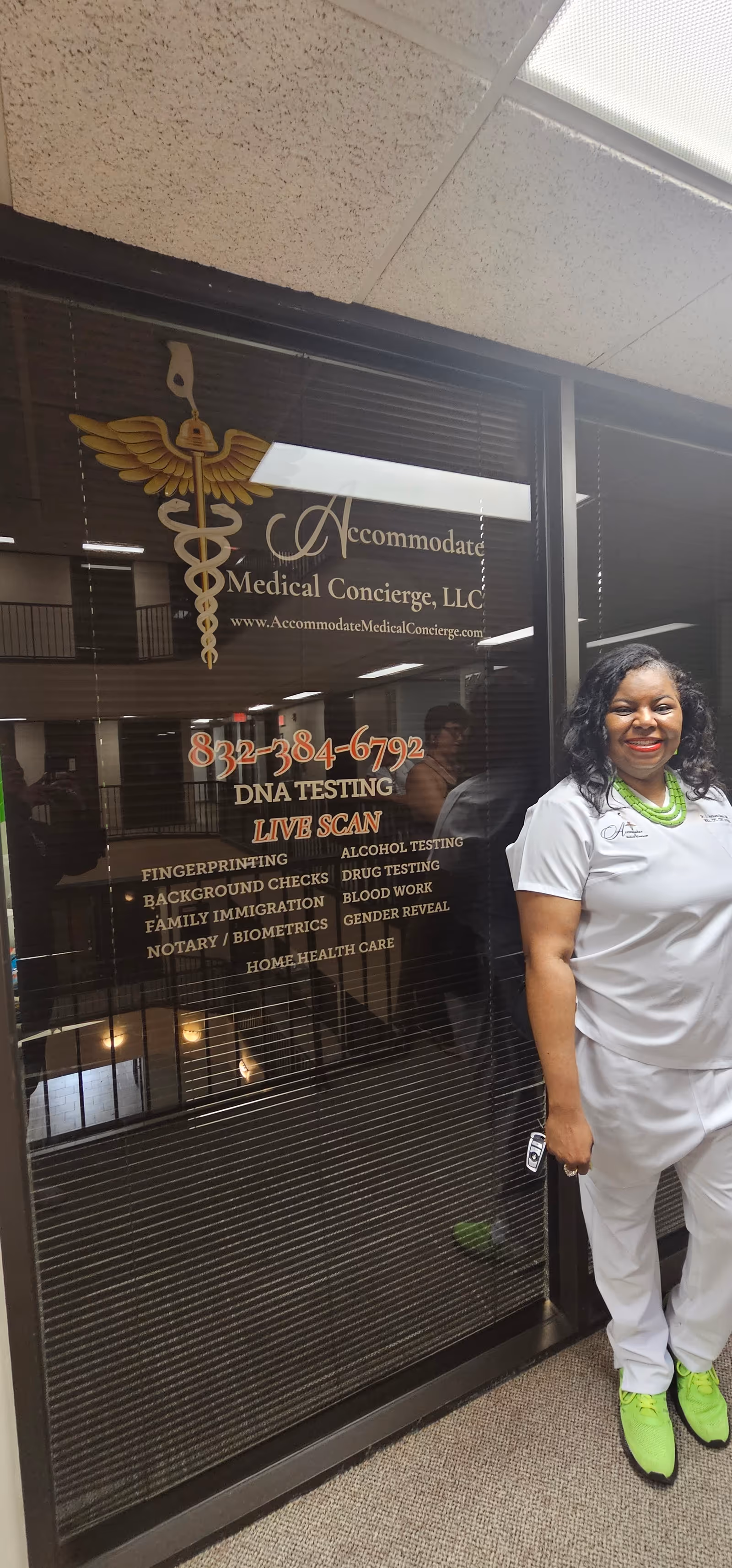 Smiling woman in scrubs stands beside glass door advertising Accommodate Medical Concierge, LLC.