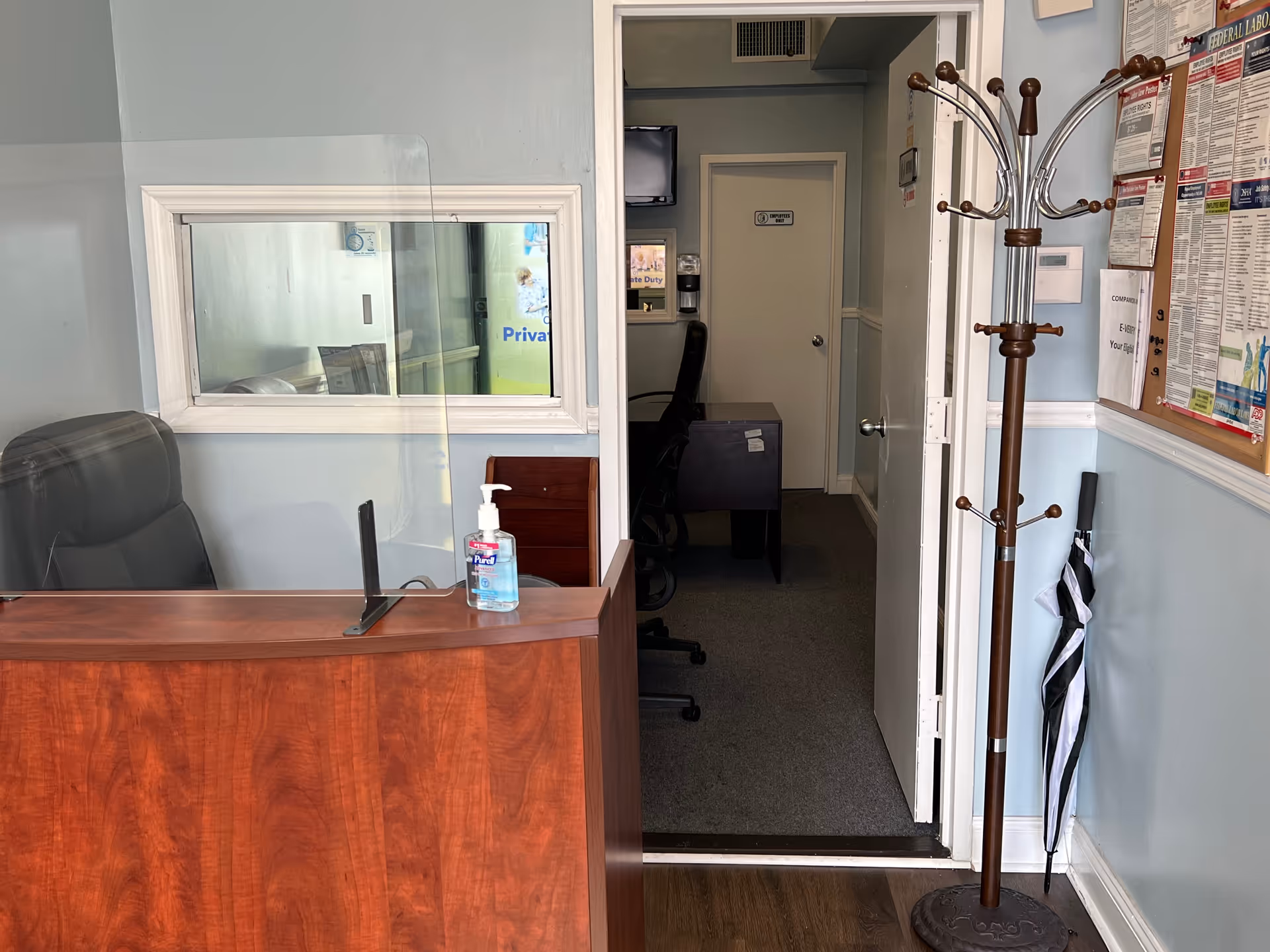 Reception desk in a hallway with chair, coat rack, sanitizer, bulletin board, and doorway to room.
