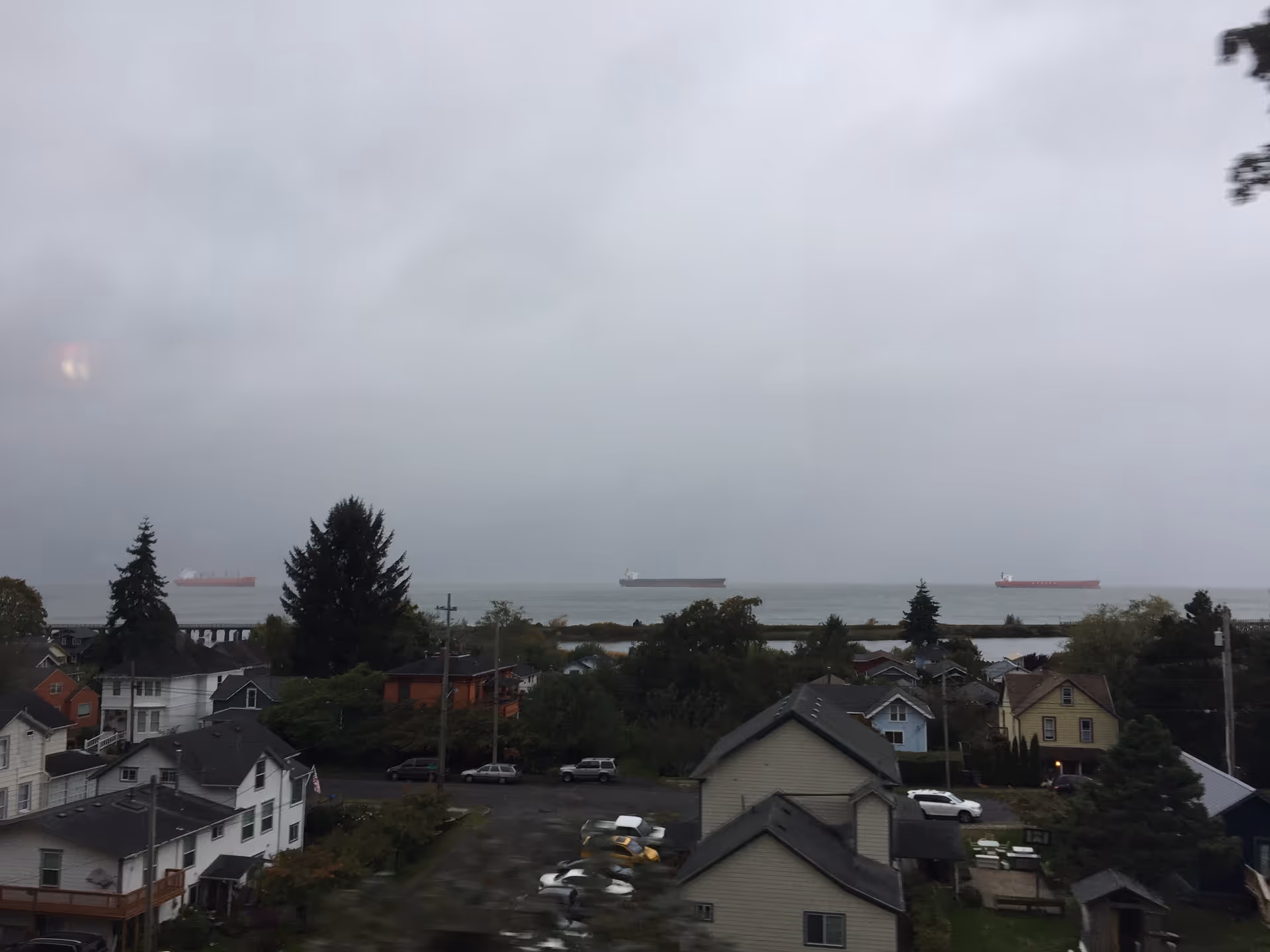 High-angle view of a coastal town with rooftops, trees, and ships on the horizon.