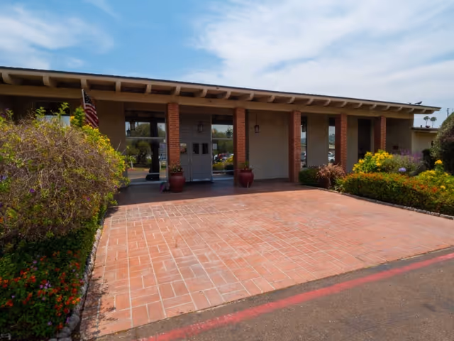Front exterior view of a single-story building with a covered entrance supported by brick columns. There are potted plants near the entrance, bushes and flowers on either side, and an American flag on the left side. The sky is clear with some clouds.