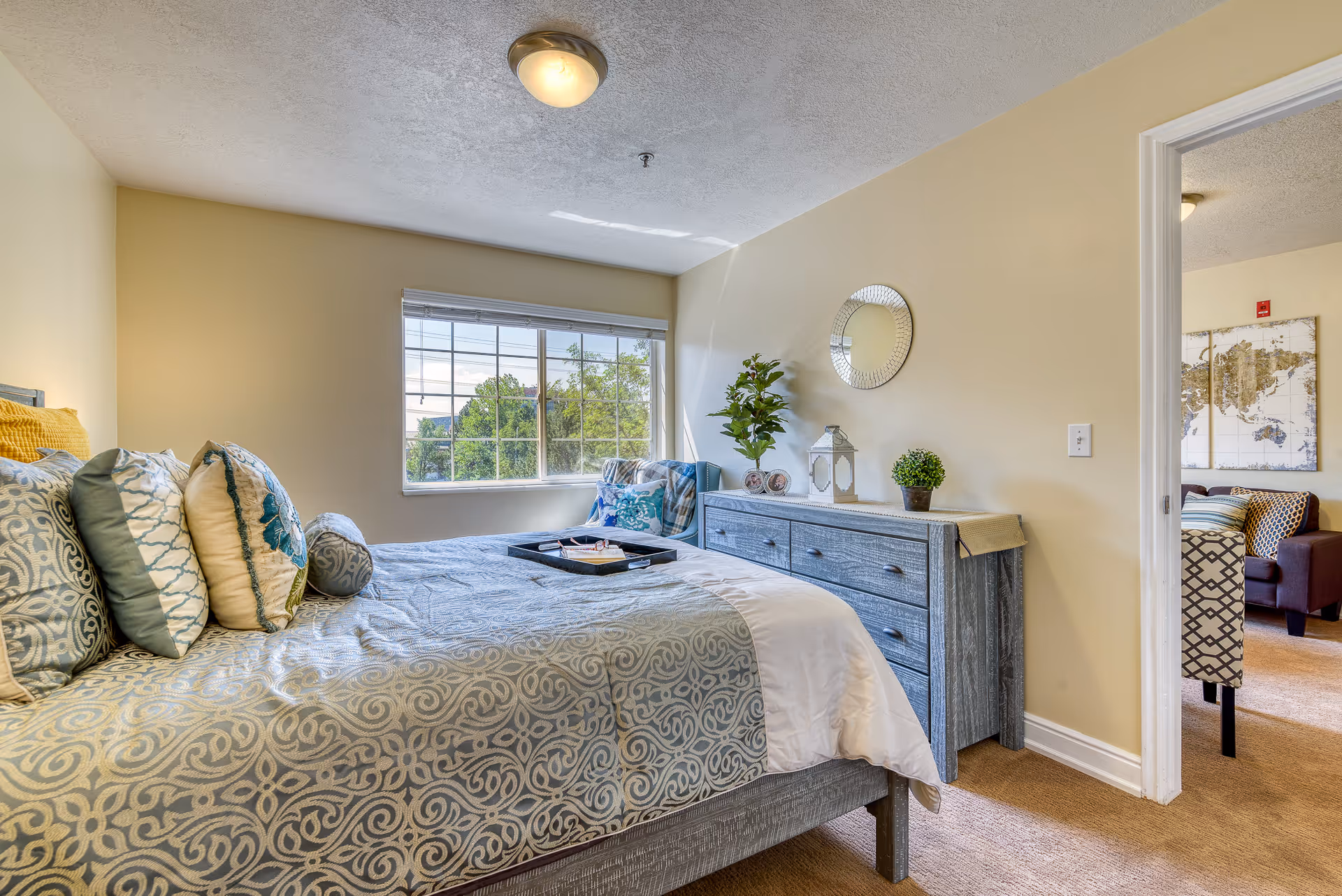 Bright bedroom with a patterned bed, dresser, window view, and doorway to another living area.