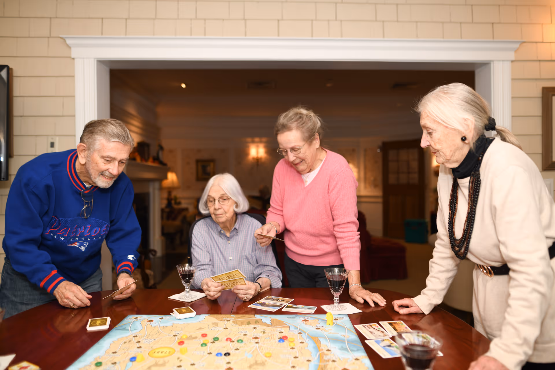 Four elderly people gathered around a wooden table playing a board game in a cozy living room setting. They appear engaged and happy, with glasses of red wine on the table. The room has warm lighting and homey decor.