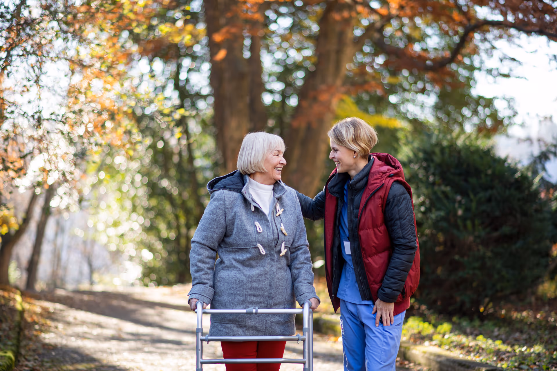 Two elderly women outdoors in a park chat; one uses a walker.