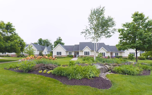 Single-story facility building with a manicured lawn, landscaped flower beds, and trees in the foreground.