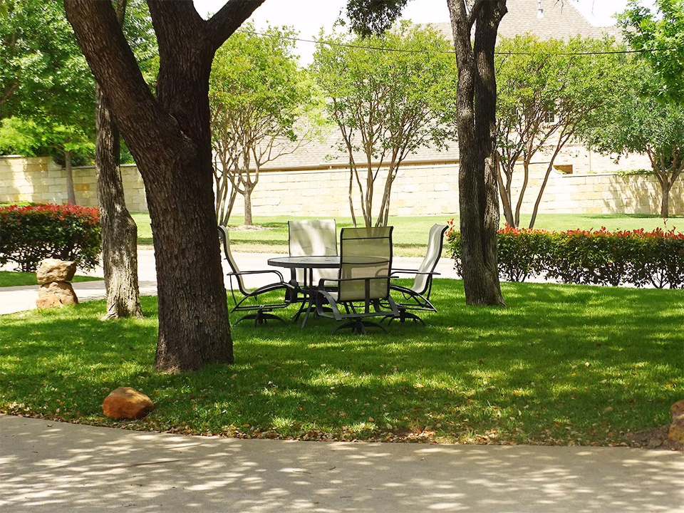 Outdoor seating area with a round table and four chairs on a grassy lawn surrounded by trees and bushes, with a stone wall and building in the background.
