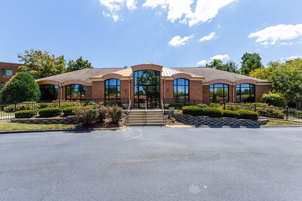 Front exterior view of a single-story brick building with large arched windows, a central glass entrance, steps leading up to the door, and landscaped bushes and trees under a blue sky with some clouds.