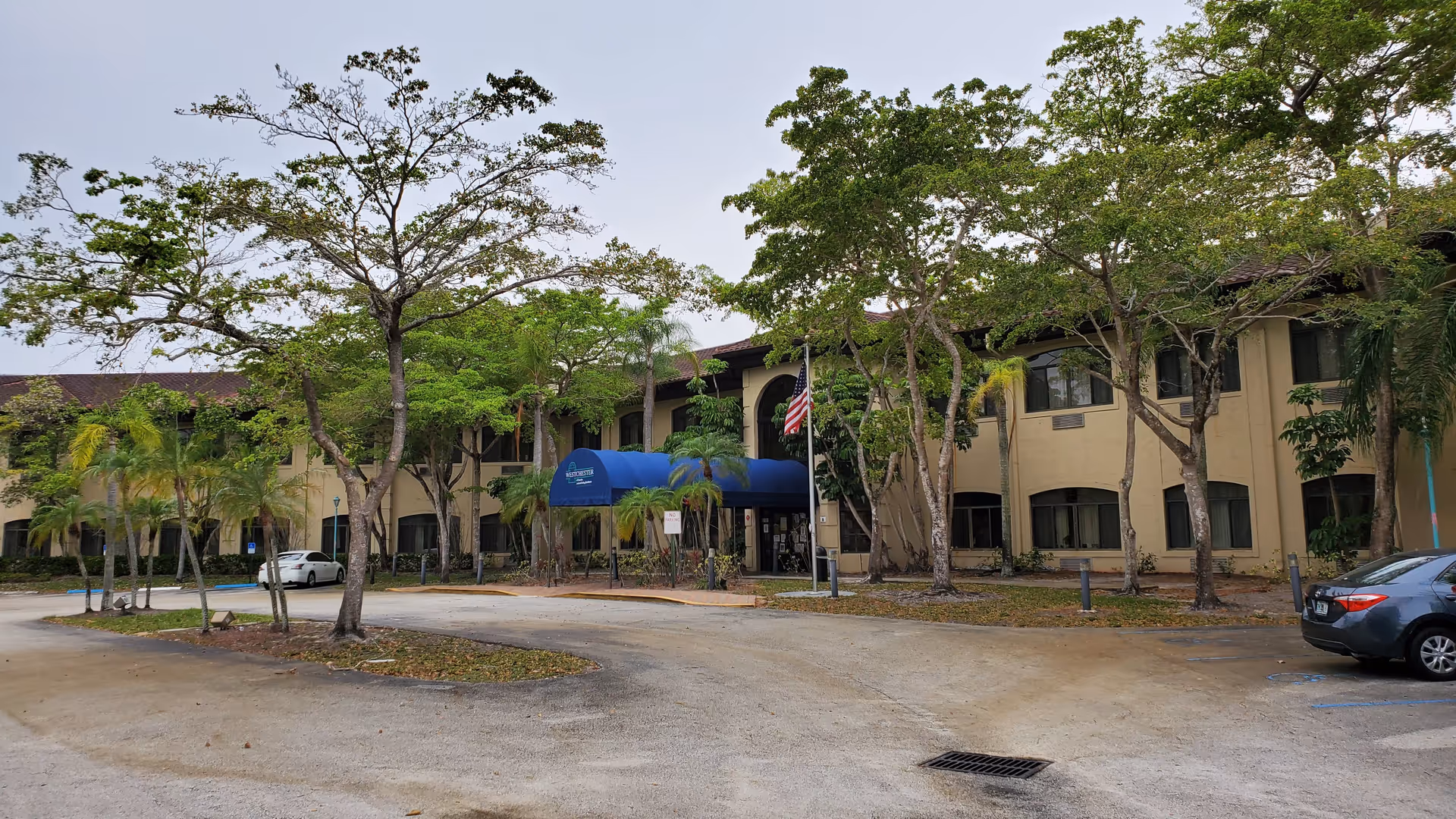 Two-story beige senior living building with a blue entrance canopy, trees, and parked cars.