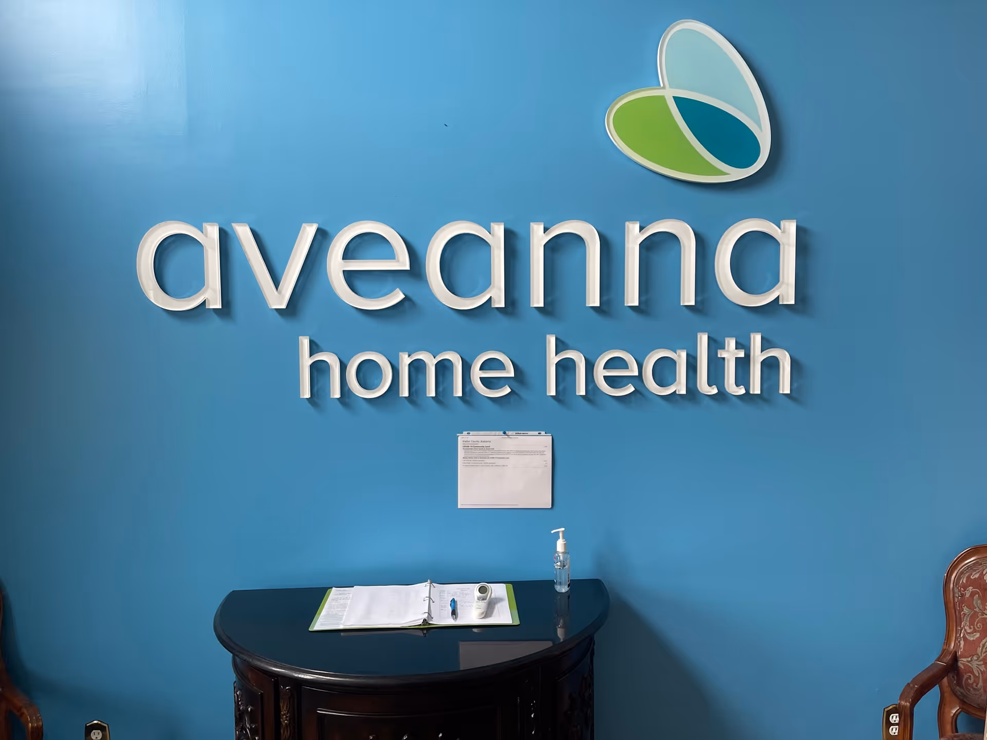 Blue wall with Aveanna Home Health logo and a reception desk with binder and sanitizer.