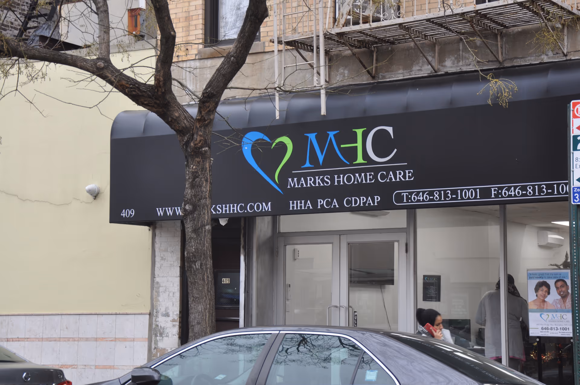 Exterior storefront for Marks Home Care with logo sign and glass entry, a tree in front.