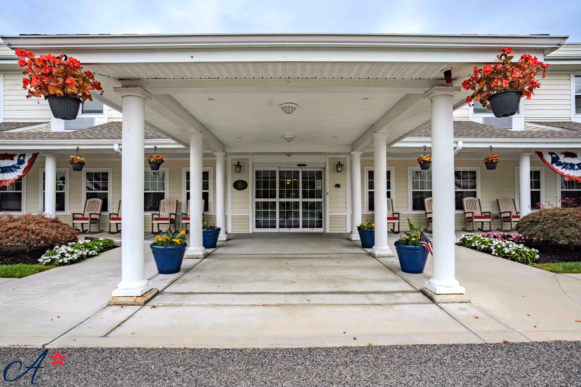 Front entrance of a senior living facility with a covered driveway supported by white columns. There are hanging flower pots with red flowers and blue planters with yellow flowers along the walkway. The building has beige siding, multiple windows, and several chairs on the porch. Patriotic bunting decorations are visible on the sides.