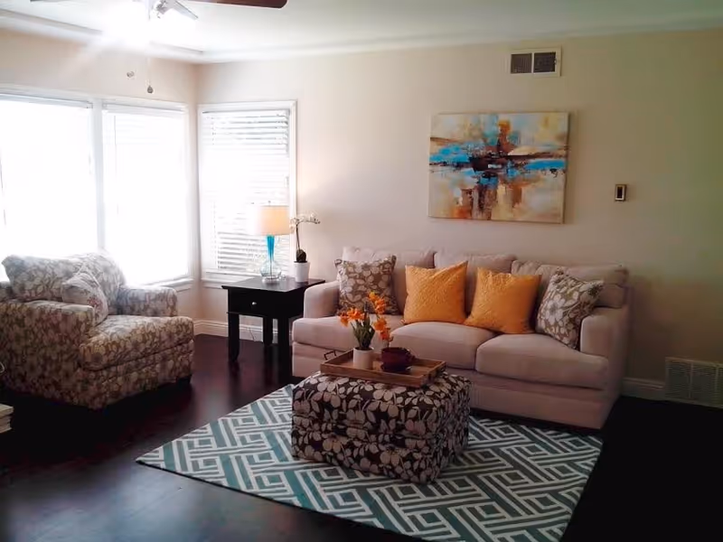 A cozy living room with a beige sofa adorned with yellow and floral cushions, a matching floral armchair, a patterned ottoman on a geometric rug, a black side table with a lamp and a small plant, and a colorful abstract painting on the wall.