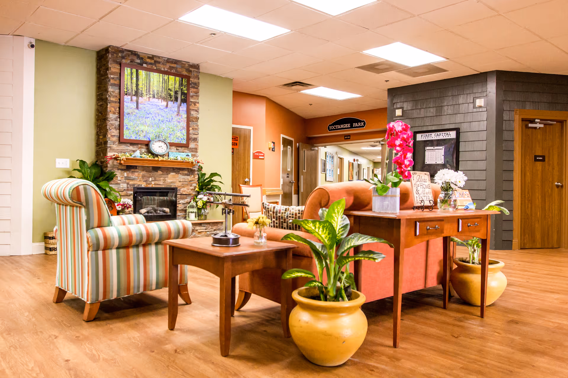 A cozy seating area in a senior living facility featuring a striped armchair, a brown sofa, wooden tables with flower vases, and large potted plants. A stone fireplace with a framed picture of a forest above it is visible on the left wall. The floor is wooden, and the ceiling has recessed lighting. Doors and a hallway are visible in the background.