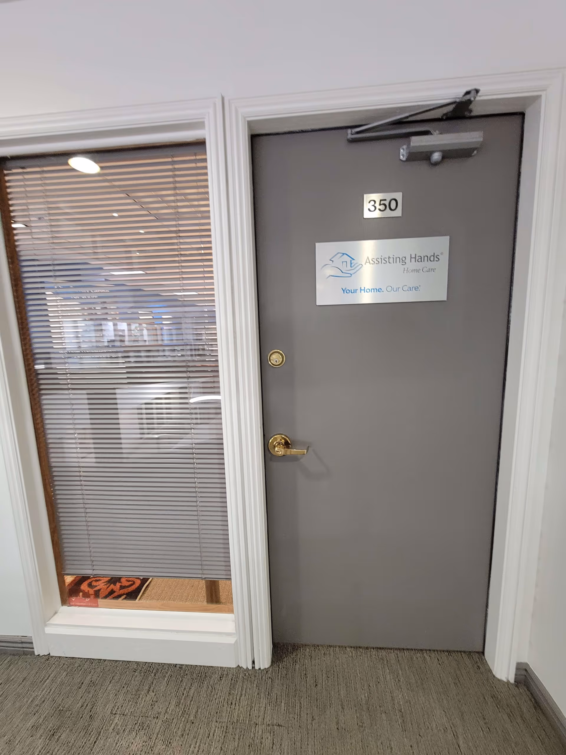 Grey door with 350 and Assisting Hands sign in a hallway beside a window with blinds.