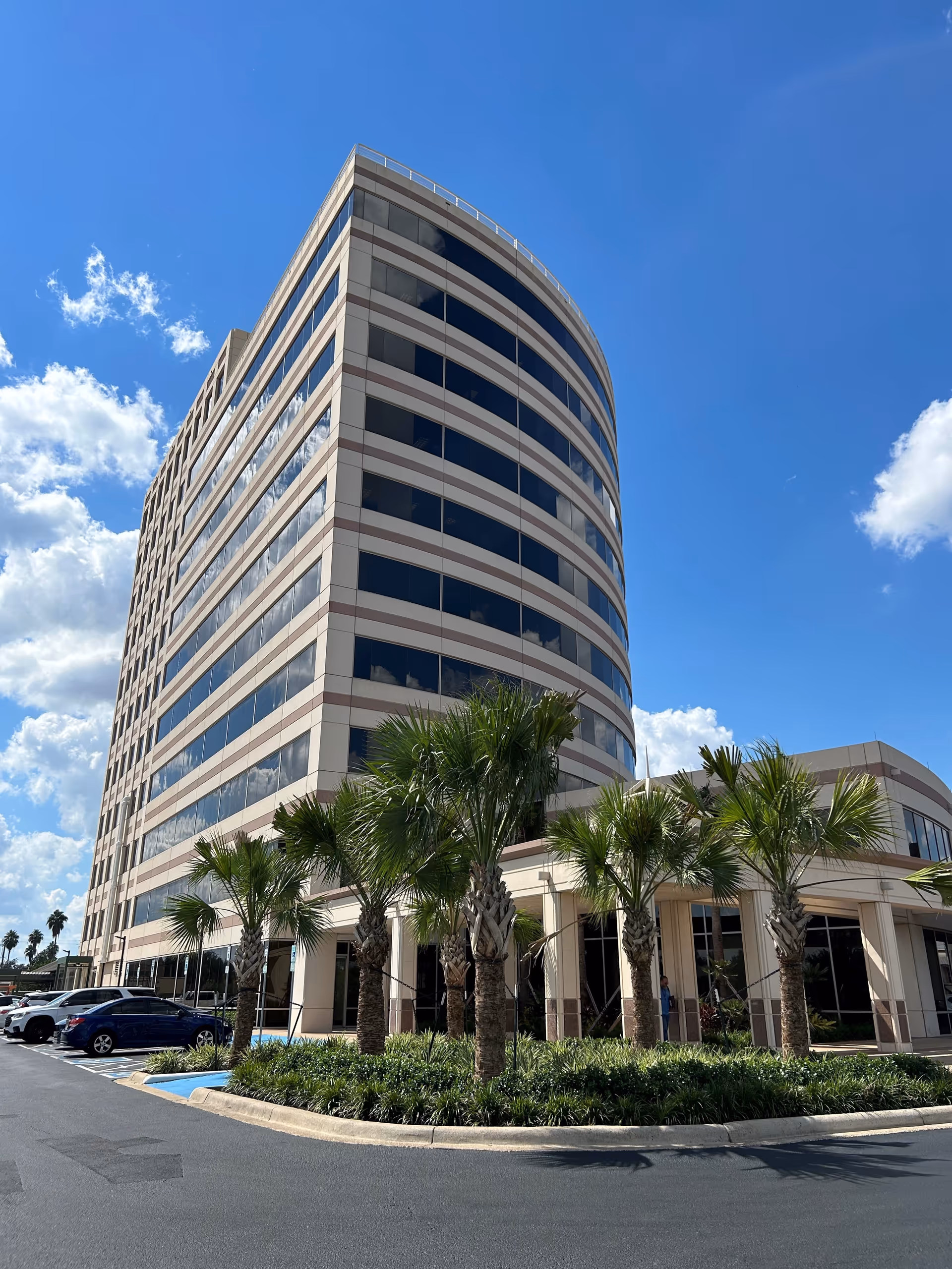 A tall curved beige office building with glass windows and palm trees in front.