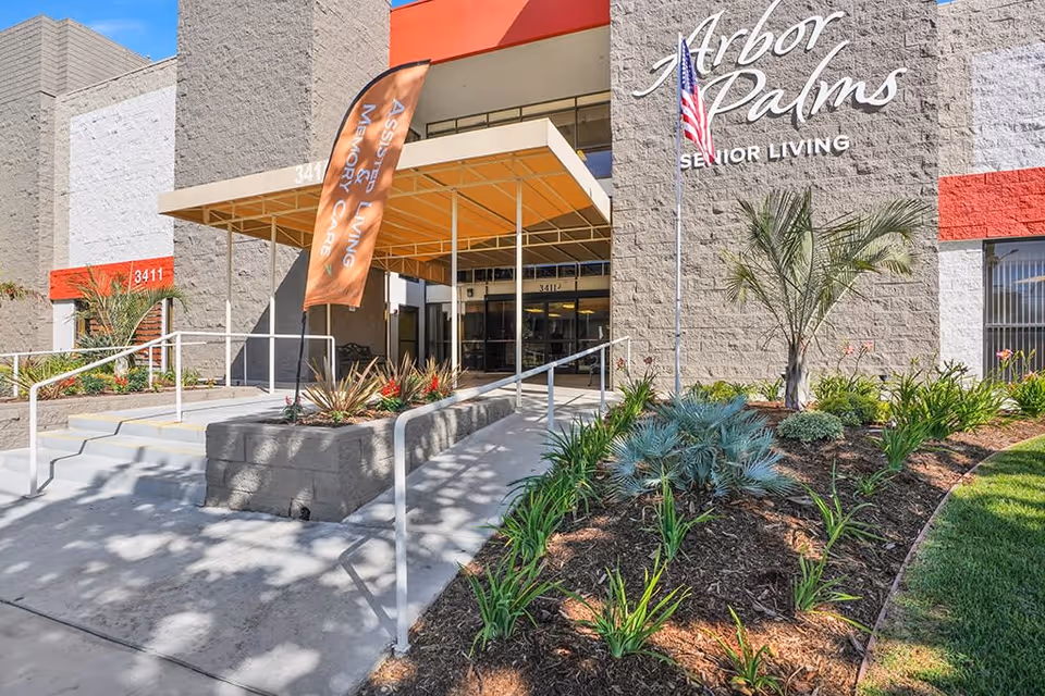 Entrance of Arbor Palms Senior Living facility with a ramp and stairs leading to the main door, a flagpole with an American flag, landscaped garden with various plants, and a banner indicating Assisted Living and Memory Care services.