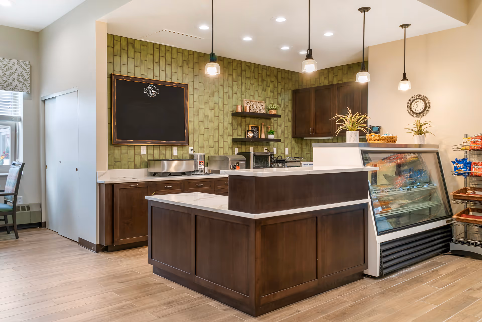 Interior kitchenette with dark wood cabinets, green tile wall, pendant lights, and refrigerated display case.