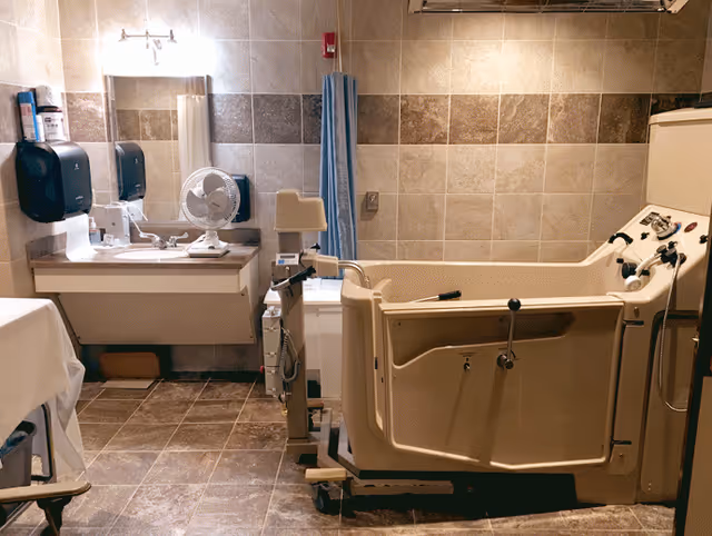 A bathroom designed for assisted bathing with a large beige walk-in bathtub equipped with multiple controls and handrails. The room has tiled walls and floor in neutral tones, a sink with a mirror above it, a small fan on the counter, and a blue privacy curtain near the bathtub.