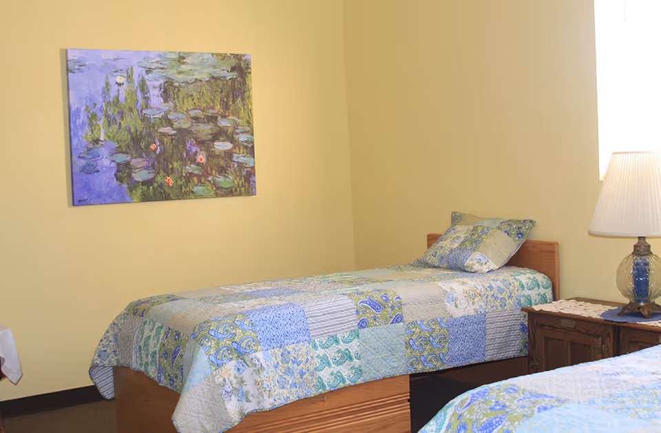 A simple bedroom with two single beds covered in patchwork quilts in shades of blue, green, and beige. A wooden nightstand with a glass lamp and a doily sits between the beds. A window with bright light is on the right wall, and a framed painting of water lilies hangs on the left wall.
