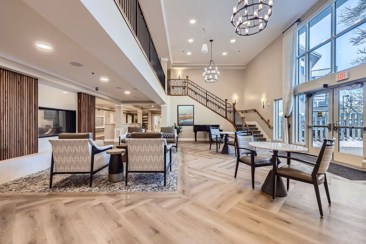 A spacious and well-lit common area in a retirement community featuring modern furniture including armchairs and round tables with chairs. The room has large windows allowing natural light to fill the space, a staircase leading to an upper level, and decorative light fixtures hanging from the ceiling. There is a fireplace with wooden paneling on one side and a piano near the staircase.