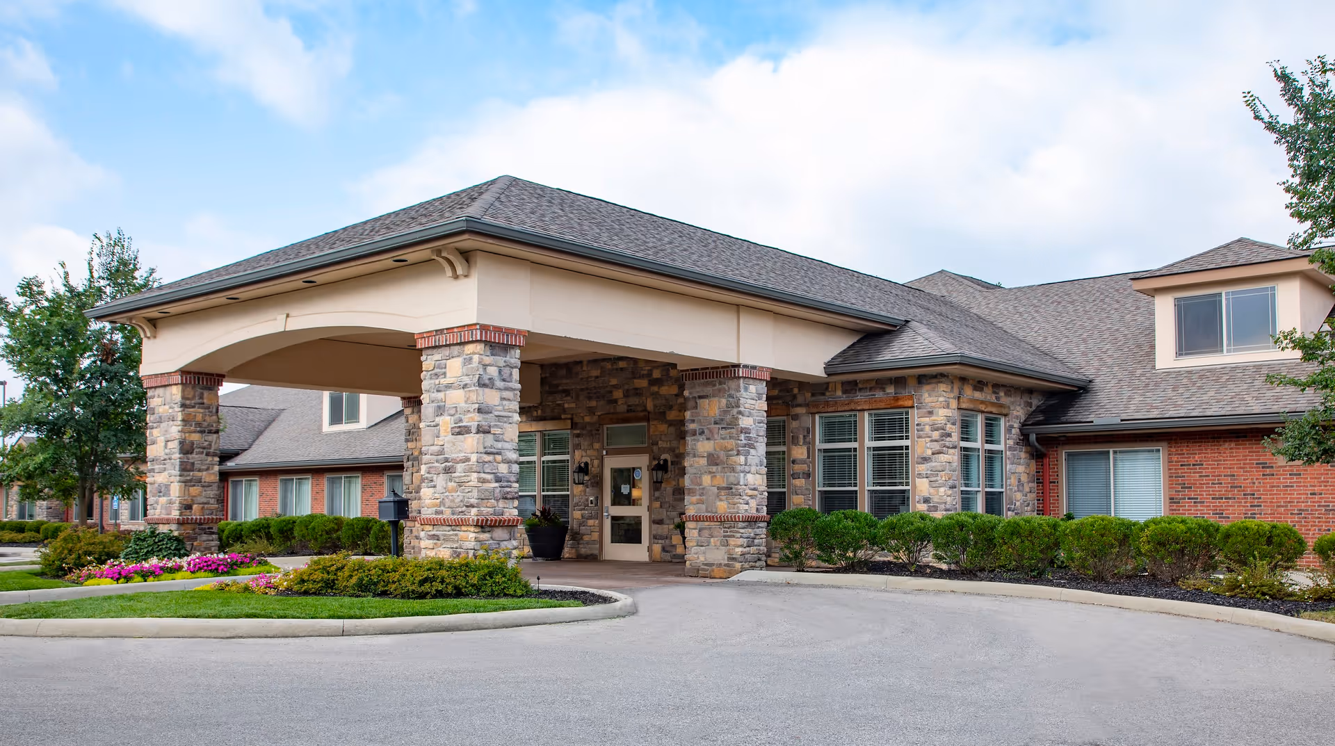 Exterior front entrance of a senior living facility with a covered driveway supported by stone pillars, surrounded by well-maintained landscaping including bushes and flowers under a partly cloudy sky.