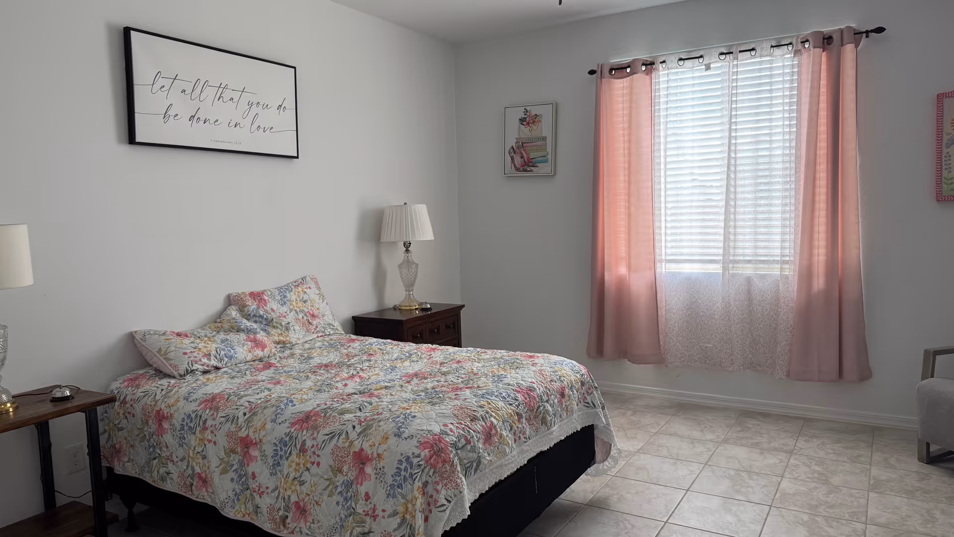 A furnished bedroom with floral bedspread, pink curtains, lamps, and two nightstands.