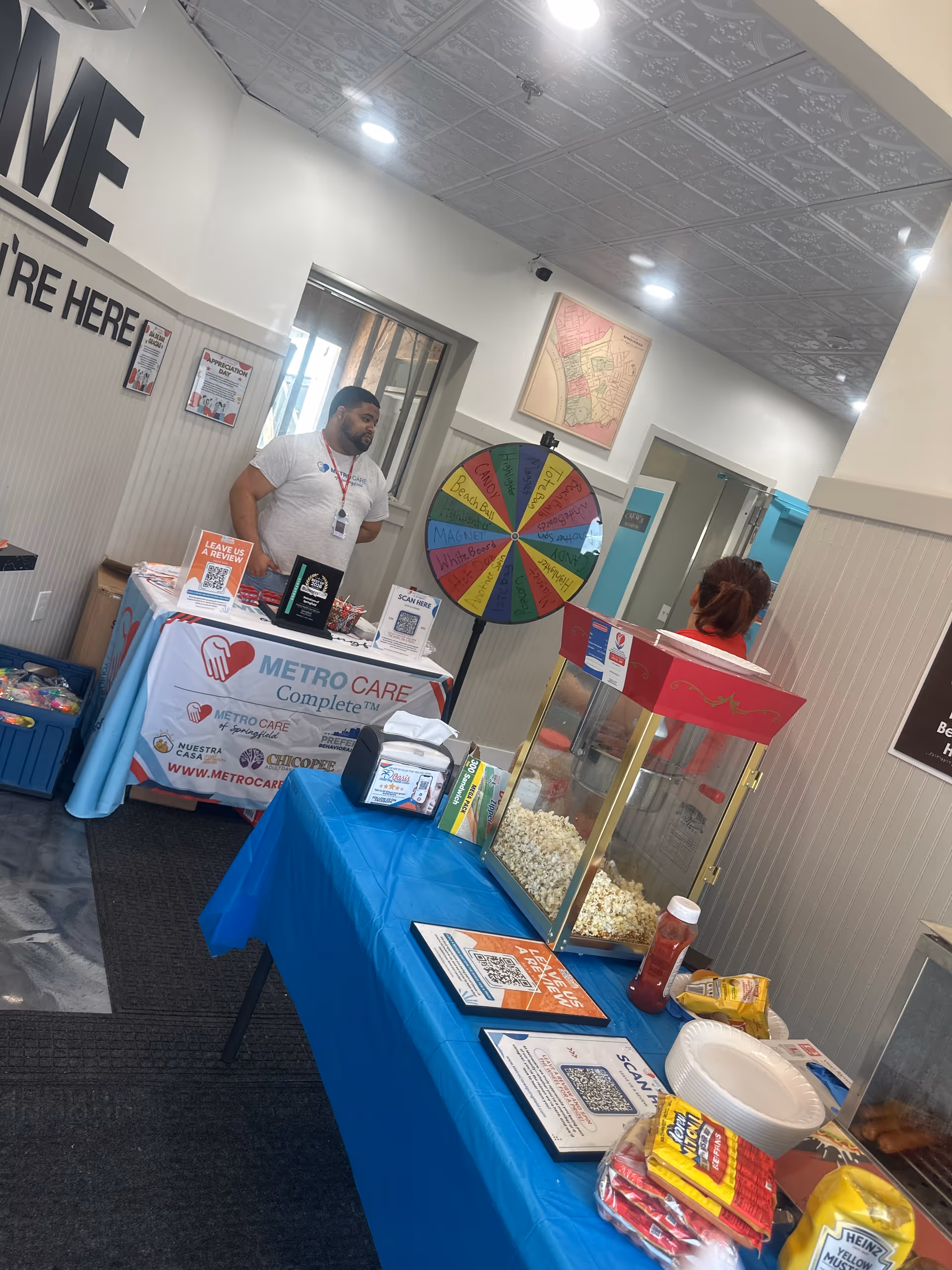 Indoor facility booth with a spinning wheel, popcorn cart, and staff at a Metro Care table.