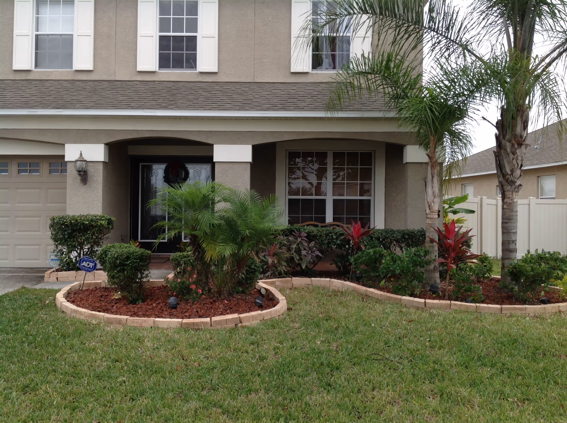 Front exterior of a two-story house with a covered porch, landscaped flower beds, and palm trees.