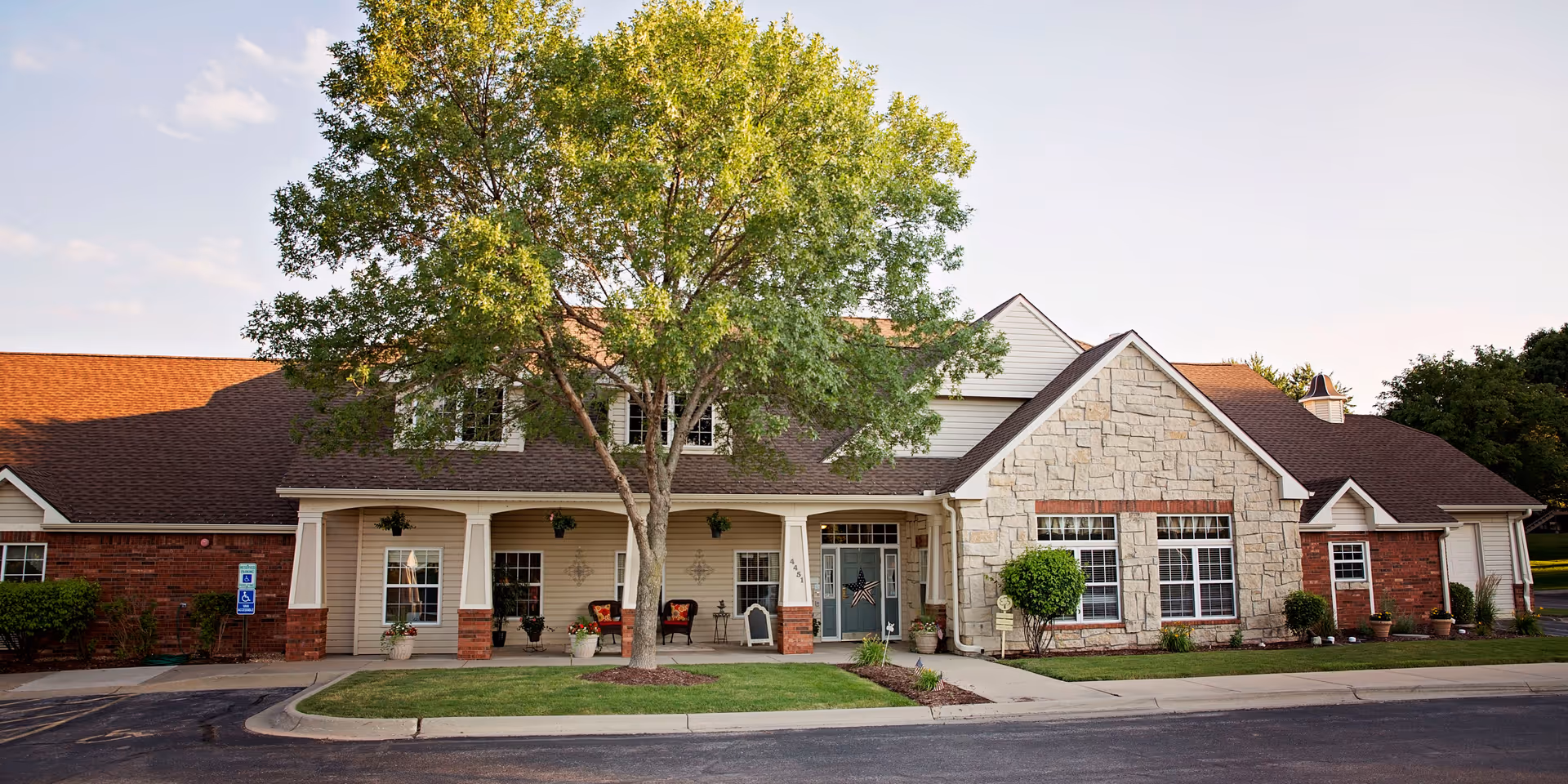 Front exterior view of Bickford of Lincoln facility with a large tree in front, a covered porch with chairs and potted plants, and a combination of brick and stone facade under a clear sky.