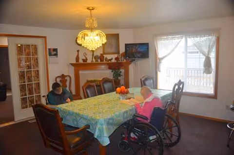 Two residents, one in a wheelchair, sit at a dining table in a senior facility.