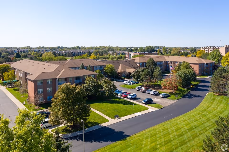 Aerial view of Brookhaven Manor senior living facility showing multiple connected brick buildings with brown roofs, surrounded by green lawns, trees, and a parking lot with several cars. The sky is clear and blue.