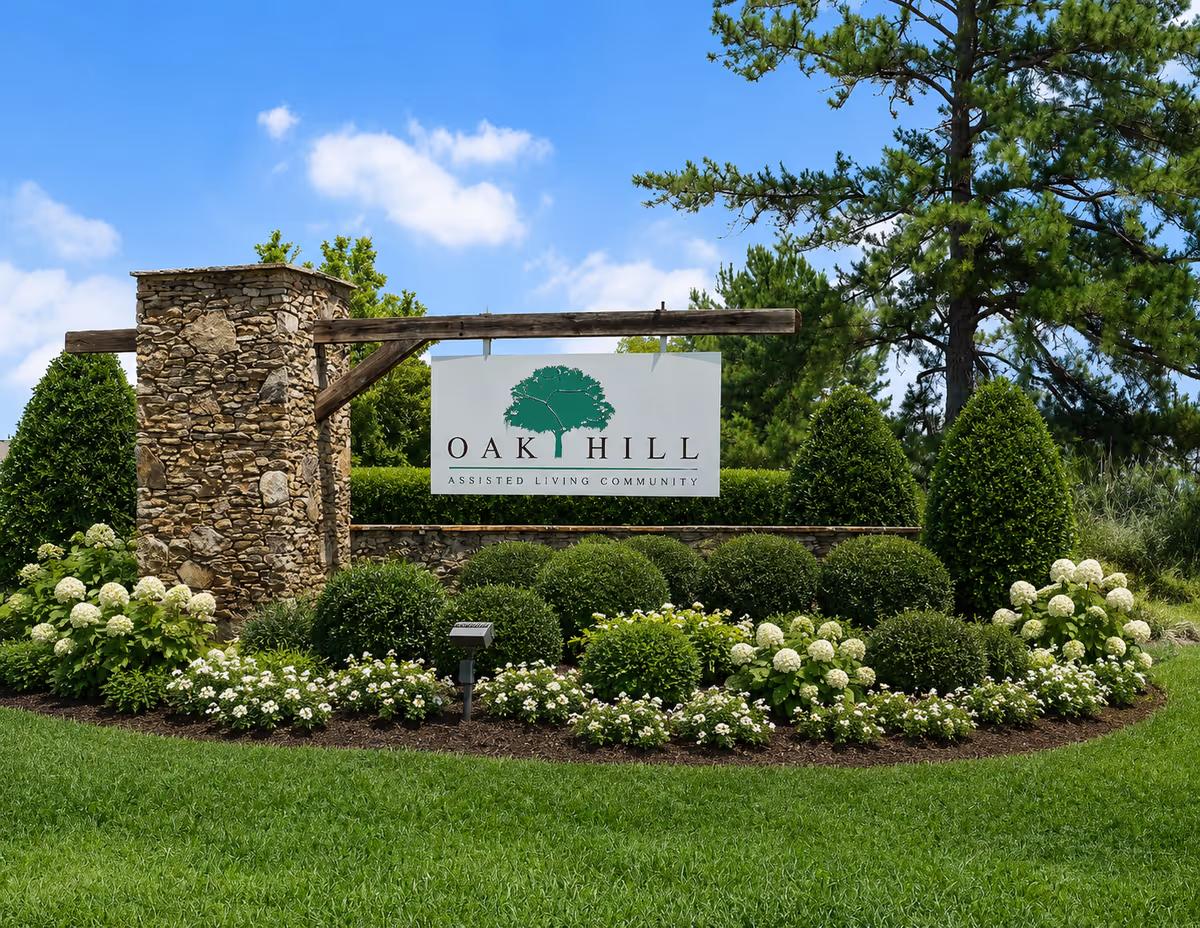 Stone entrance sign reads Oak Hill Assisted Living Community surrounded by trimmed shrubs and flowers.