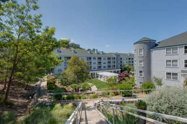 Outdoor courtyard and landscaped grounds of a multi-story senior living building with a wooden walkway leading down to a central lawn and surrounding apartments.