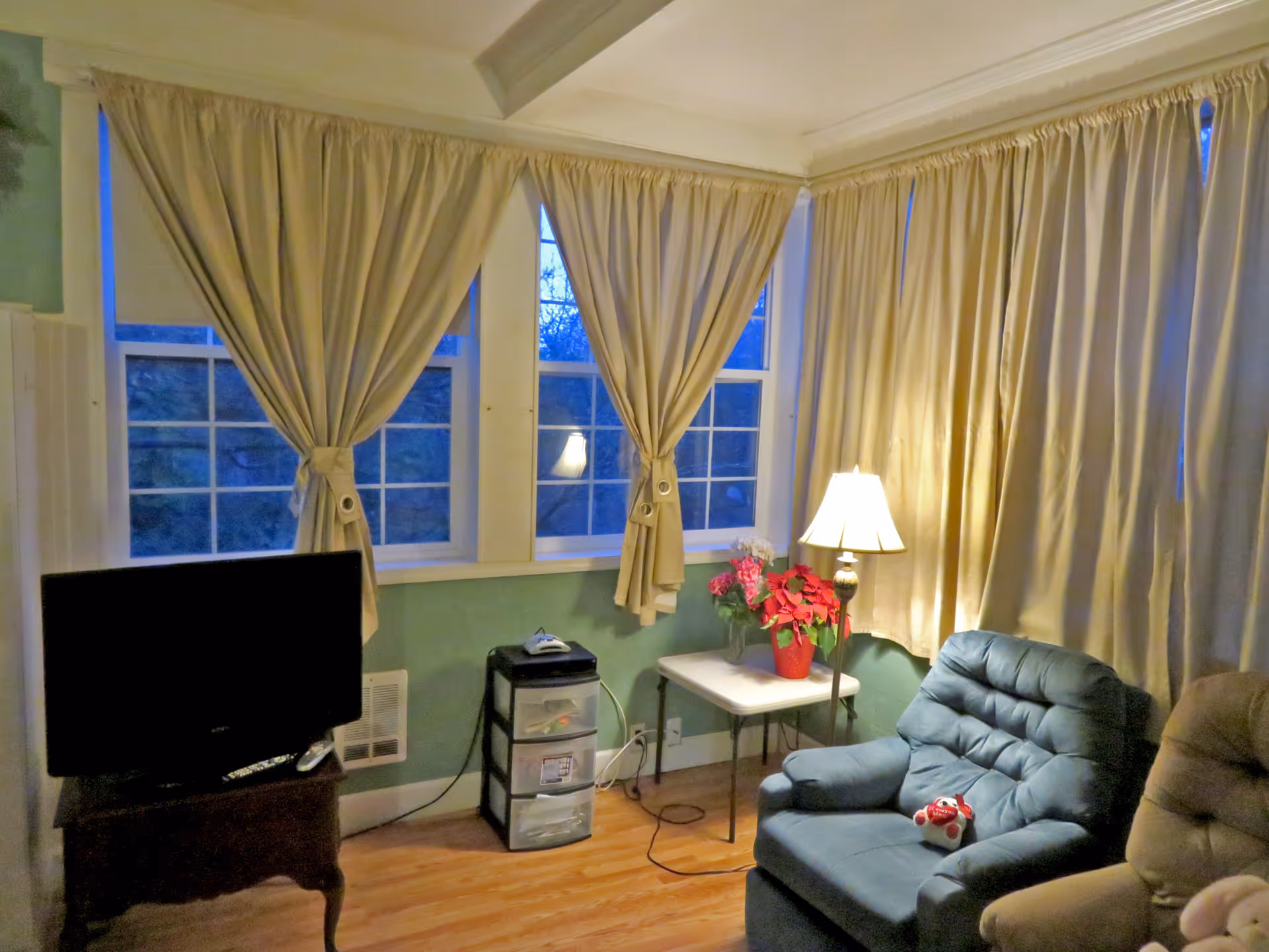 Cozy living room with large windows, beige curtains, sofa, TV, and side table with lamp.
