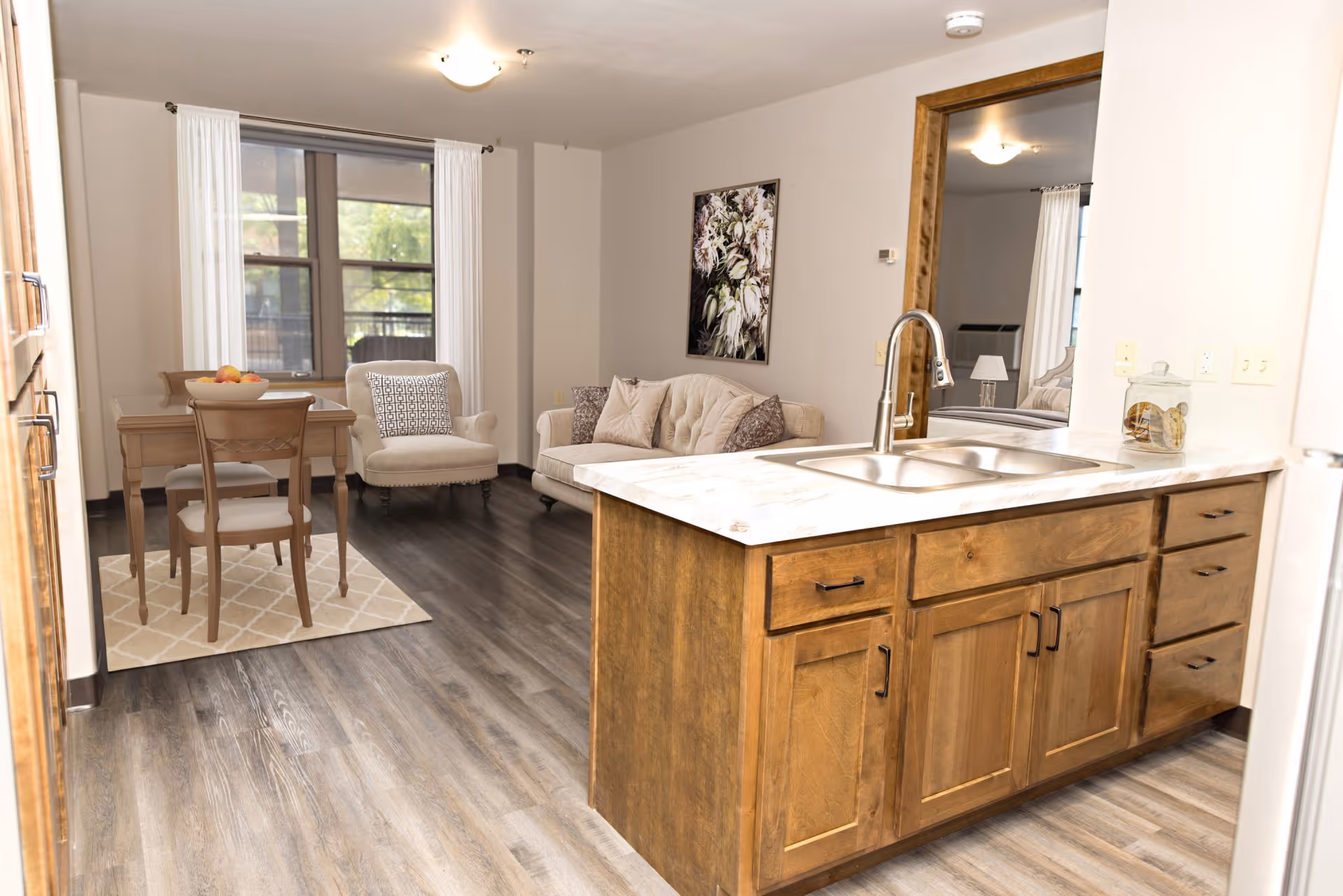 Interior view of a senior living apartment featuring a kitchen island with a sink and wooden cabinets in the foreground. In the background, there is a small dining area with a wooden table and chairs on a patterned rug, a beige armchair, and a beige sofa with cushions. A large window with white curtains lets in natural light, and a floral painting hangs on the wall above the sofa.