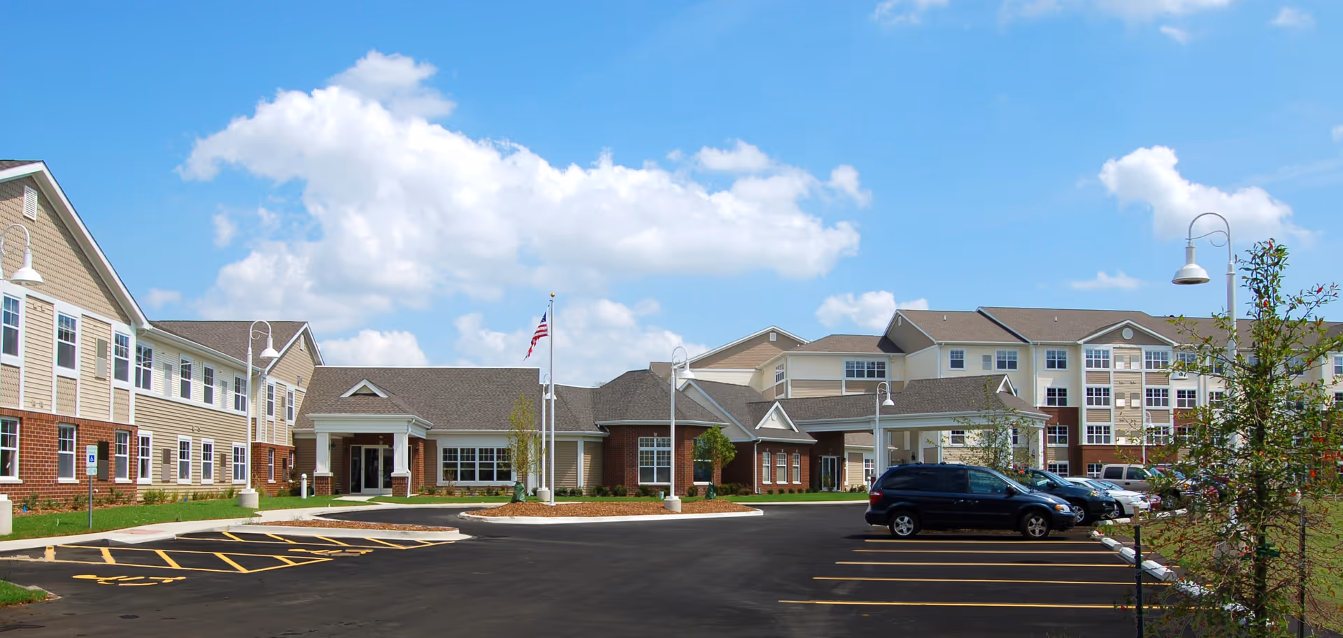 Exterior front view of a multi-story senior living facility with a parking lot, American flag, and blue sky.