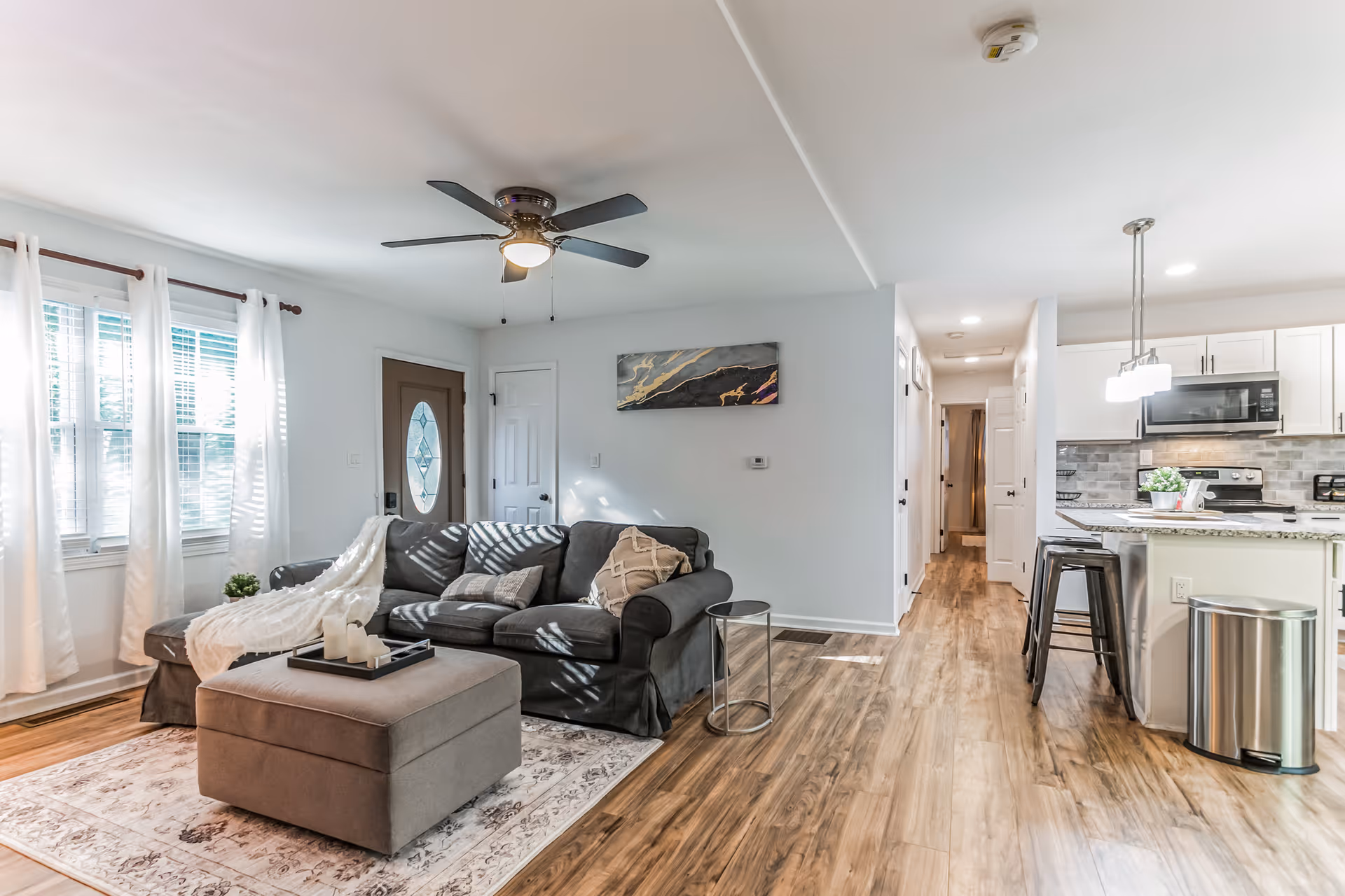 Bright open living room with a gray sectional, ottoman, rug, and adjacent kitchen.