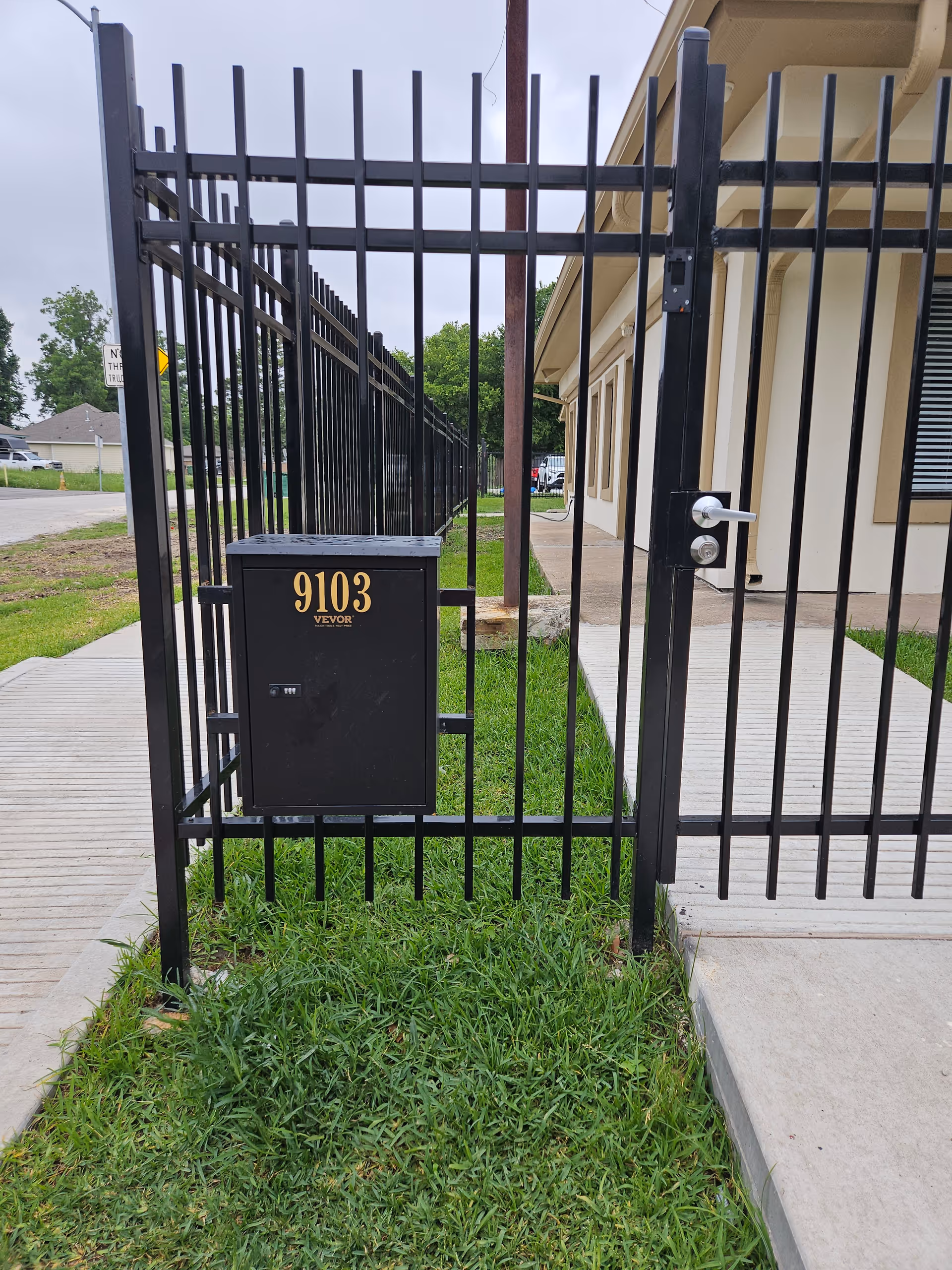 Black metal fence and gate beside a beige building with a utility box and grass.