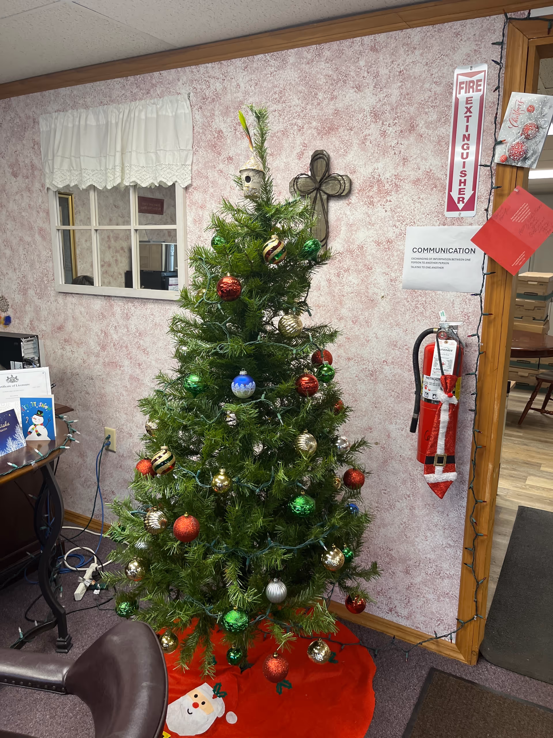Indoor Christmas tree with ornaments and string lights beside pink wallpaper and a fire extinguisher.