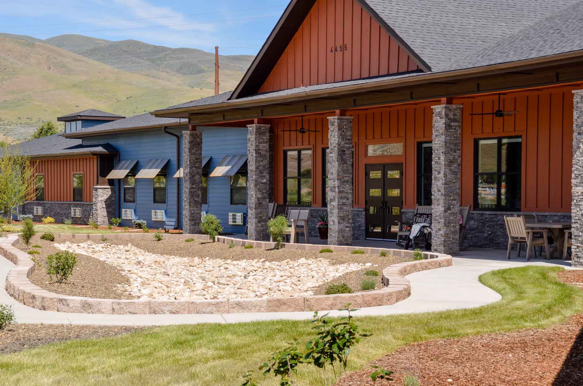 Exterior view of Aspen Valley Senior Living facility showing a building with a covered porch supported by stone columns, outdoor seating, and a landscaped area with a dry rock bed and green grass. Hills are visible in the background under a clear sky.