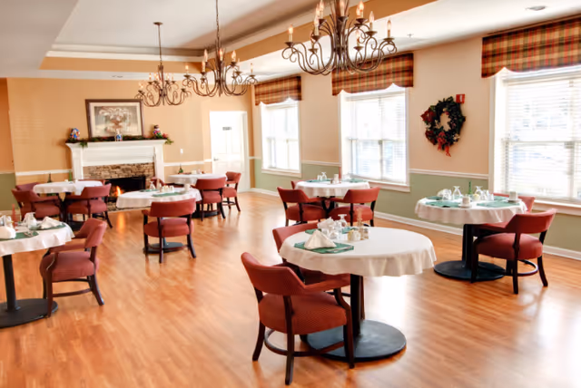 Bright senior living dining room with round tables, red chairs, and a fireplace.