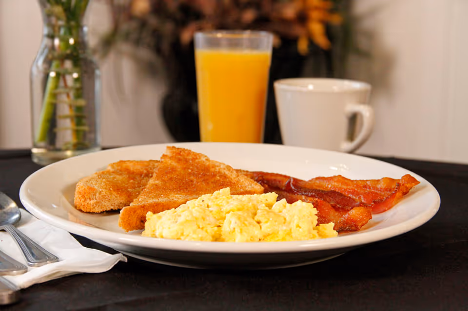 Plate of scrambled eggs, toast, bacon, and orange juice on a dining table.