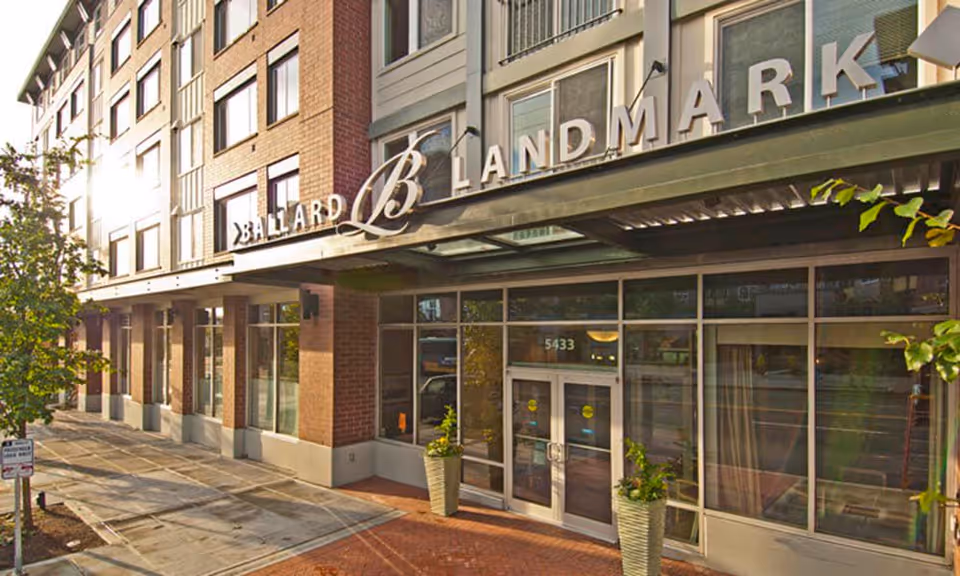 Exterior view of the Ballard Landmark building entrance with large glass doors, brick and concrete facade, and the name 'Ballard Landmark' displayed prominently above the entrance. Two large potted plants flank the doorway and a tree is visible on the sidewalk.